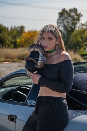 A figure in black athletic wear poses beside a car while holding a cat in an outdoor setting.