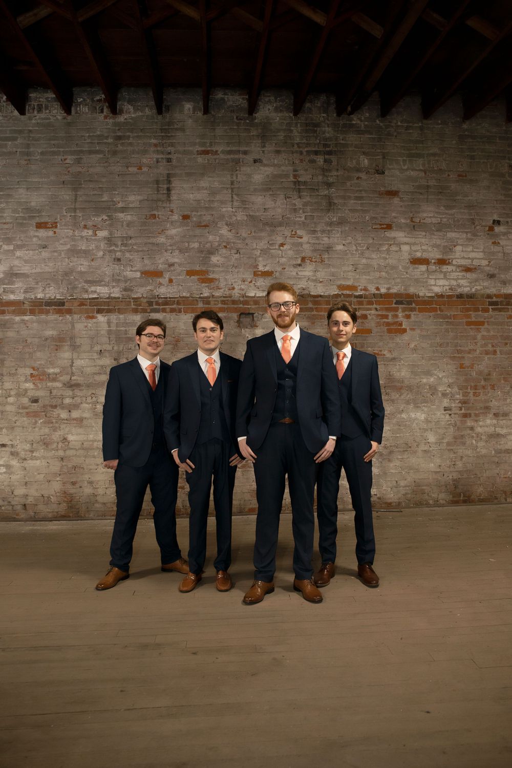 Four groomsmen in matching navy suits pose against an industrial brick wall backdrop.