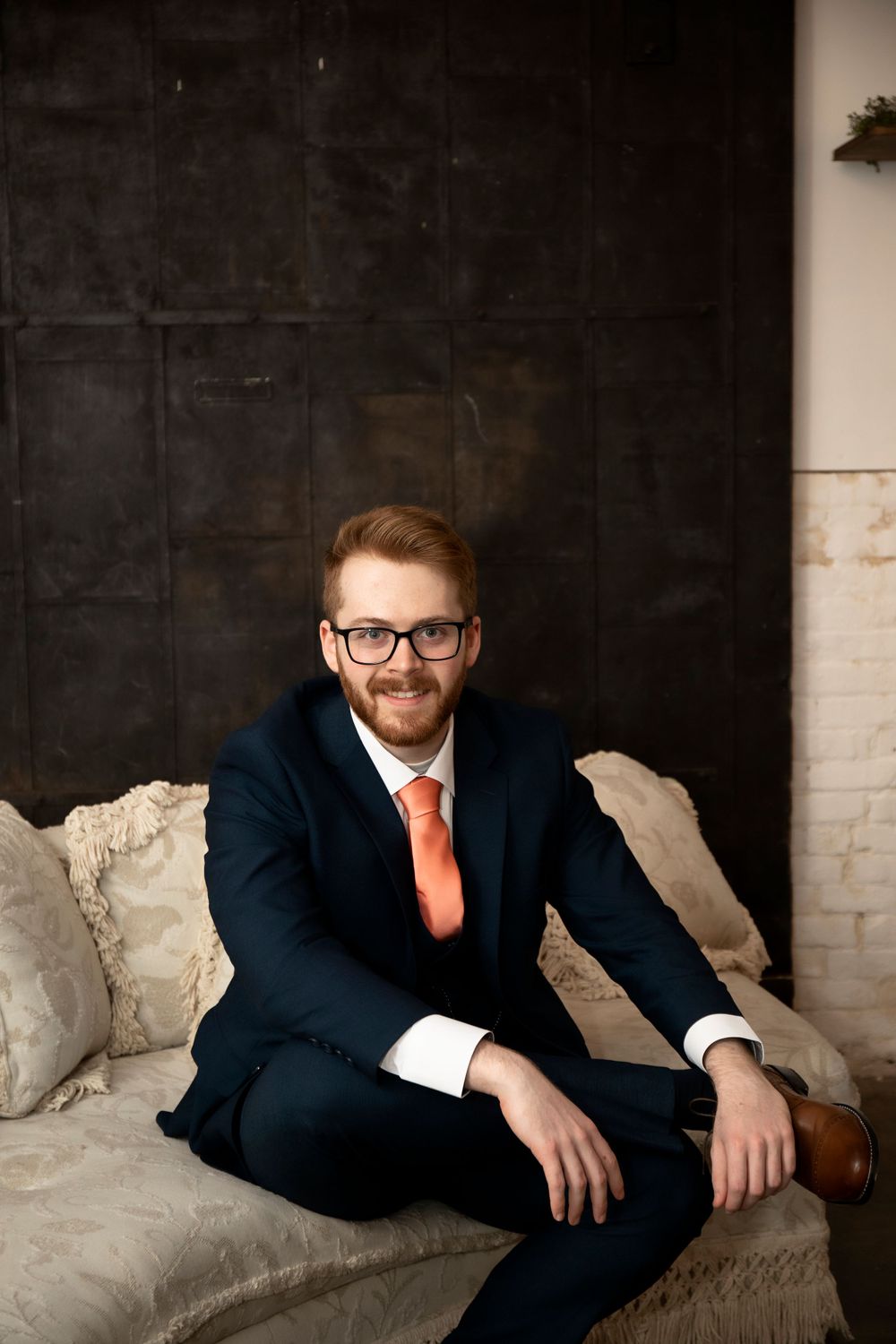A formal portrait of a person in a navy suit with orange tie seated on a couch against a dark wall.