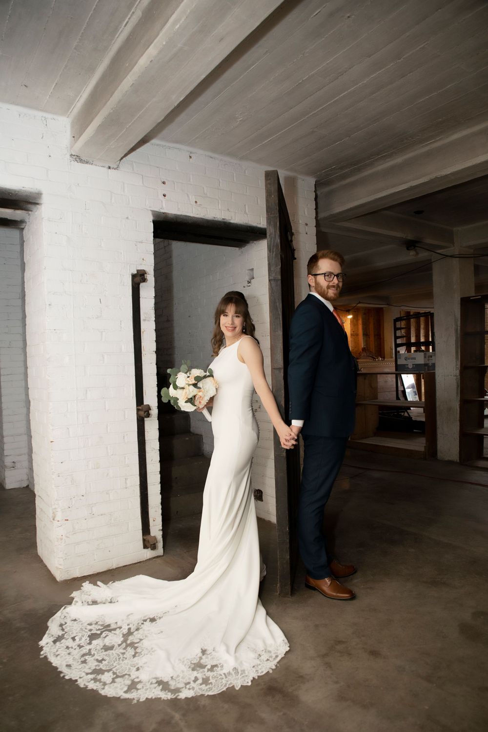 A wedding couple poses back-to-back in an industrial space with white walls and concrete floors.