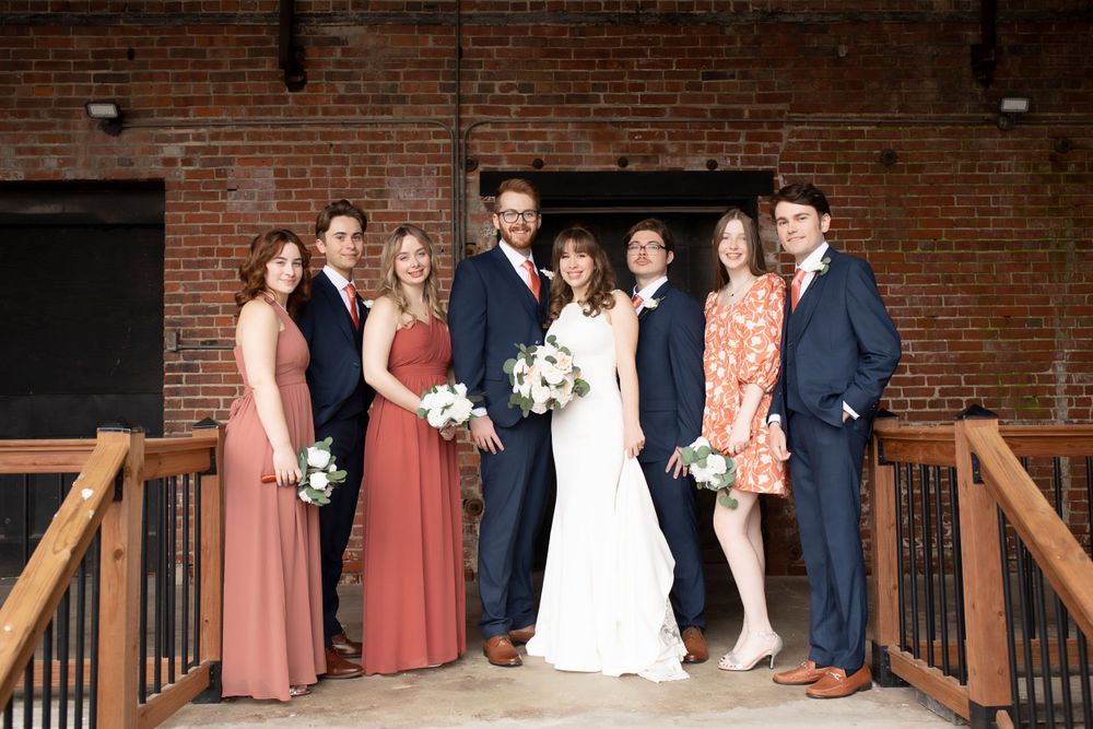 Wedding party poses together on wooden deck against brick wall backdrop.