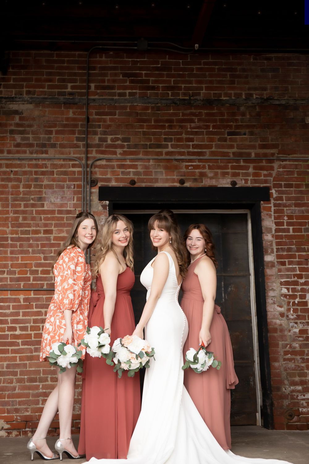 Bridesmaids in coral and floral dresses pose with bride against rustic brick wall.