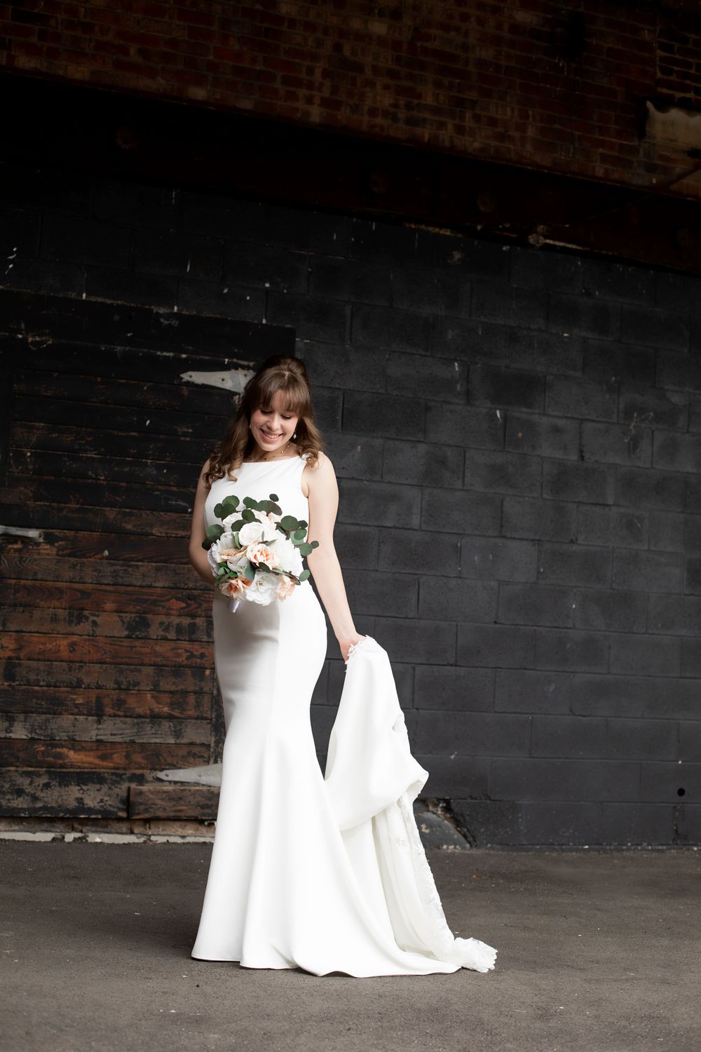 Bride in fitted white gown holds bouquet while posing against dark industrial wall.
