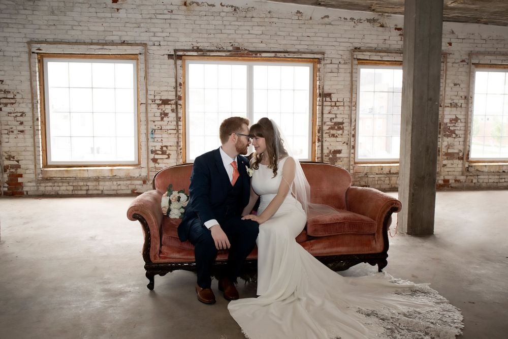 A wedding couple sits together on a vintage leather couch in a rustic brick-walled room.