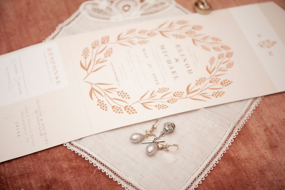 Elegant wedding invitation with rose gold floral wreath design and pearl earrings on pink fabric.