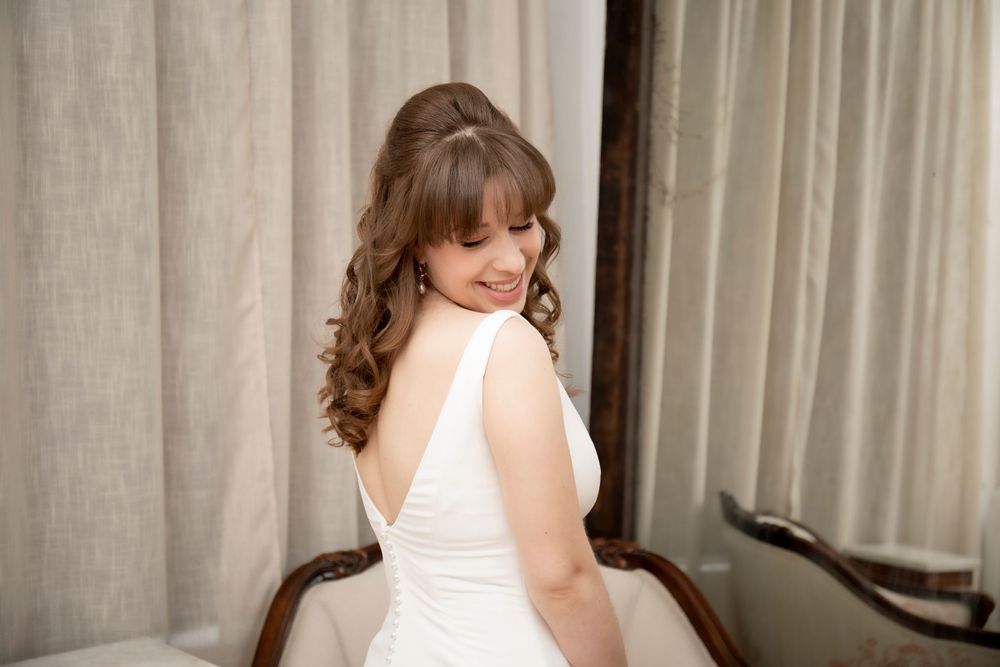 Bride in classic white dress shows off elegant back detail against soft curtained background.