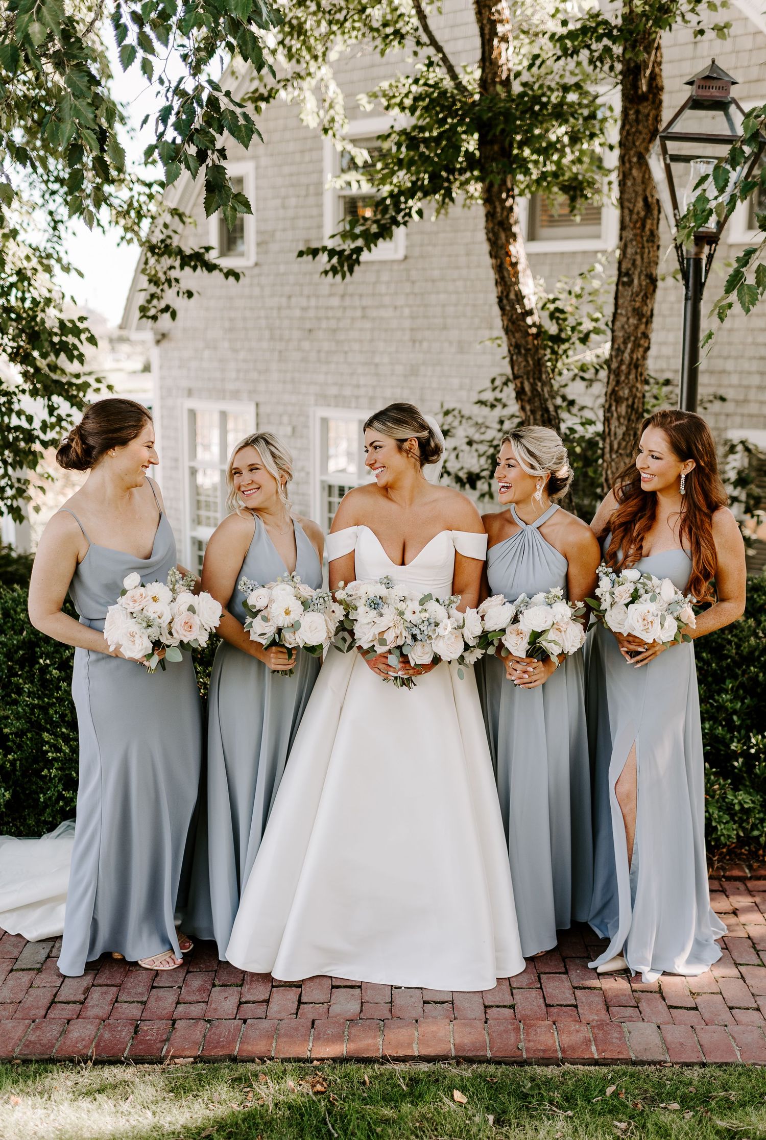 Bridesmaids in flowing grey dresses hold white bouquets while posing together on brick pathway.