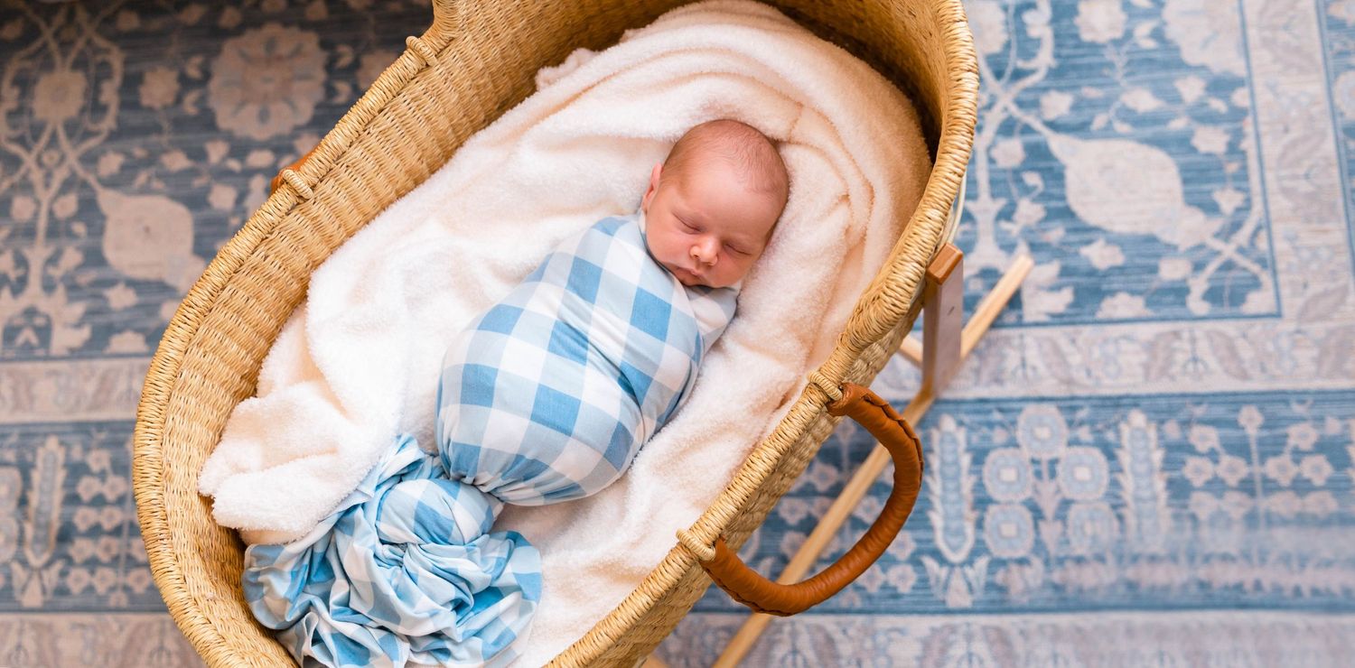 A sleeping baby in blue gingham outfit resting in a woven bassinet.