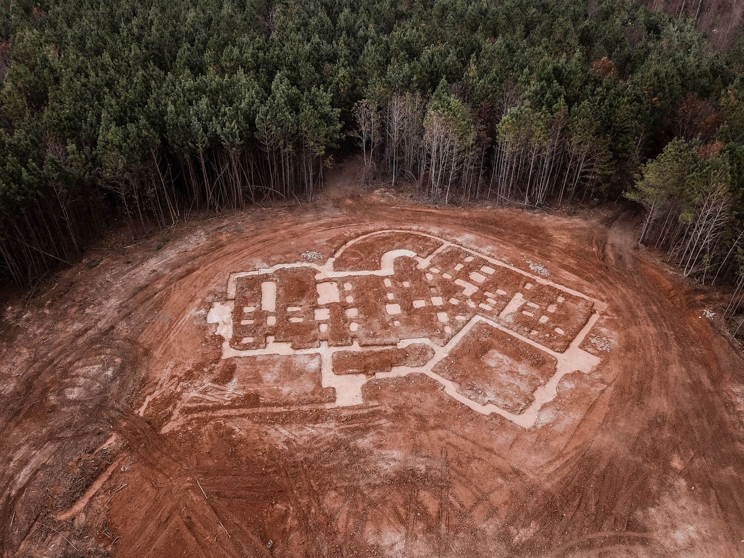 Drone shot of foundation layout marked in white on red clay soil, surrounded by forest.