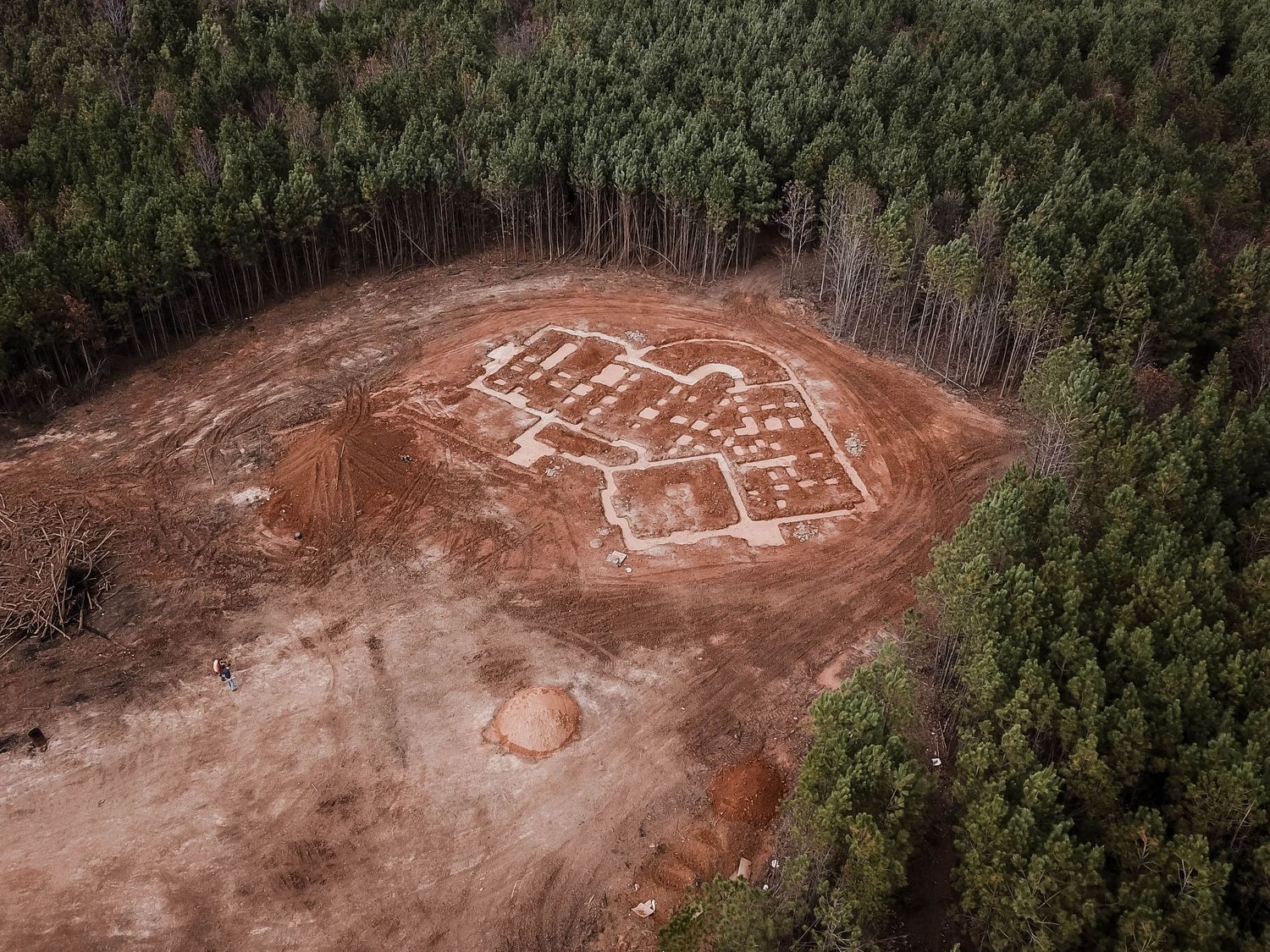 Bird's eye view of house foundation markings on red soil surrounded by green forest.