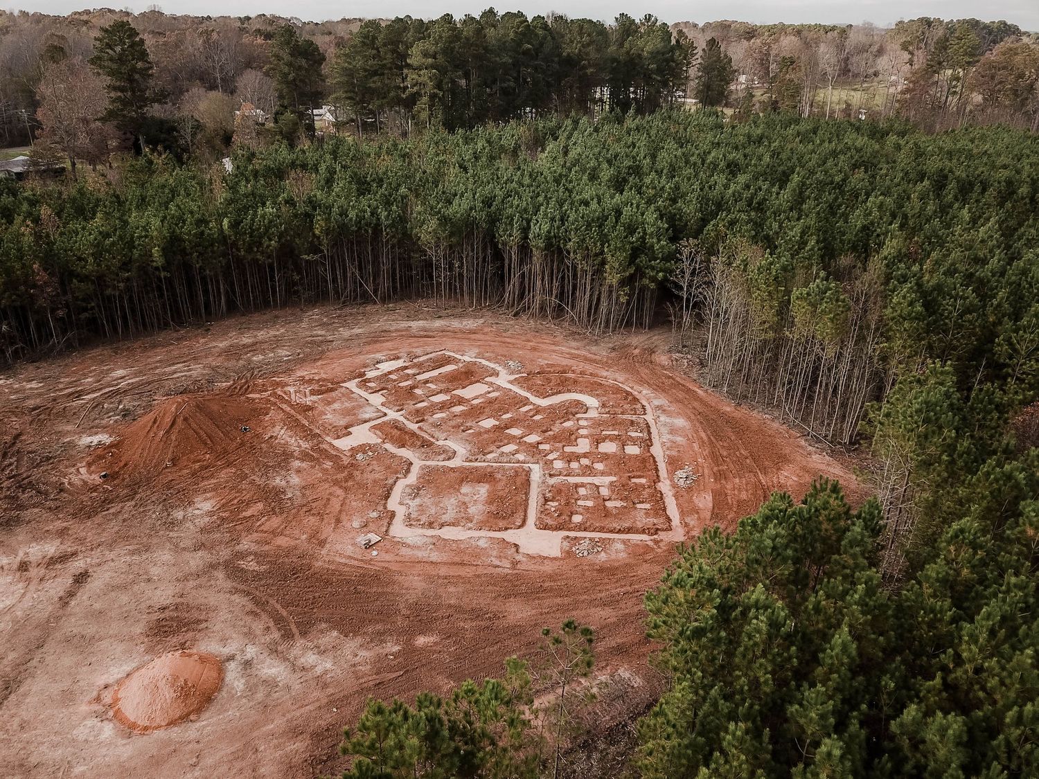 Drone shot of building foundation layout on cleared red dirt surrounded by dense pine forest.