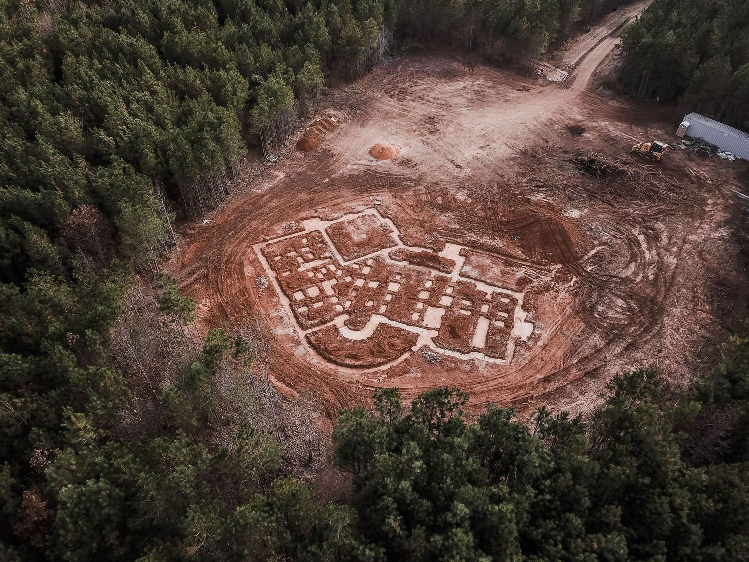 Aerial view of a cleared construction site showing foundation outline in red clay soil surrounded by forest.