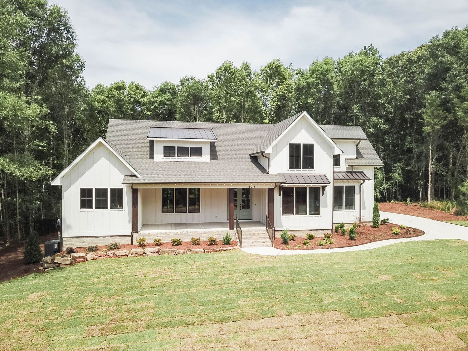 Contemporary white farmhouse with covered front porch and dormer windows surrounded by wooded landscape.