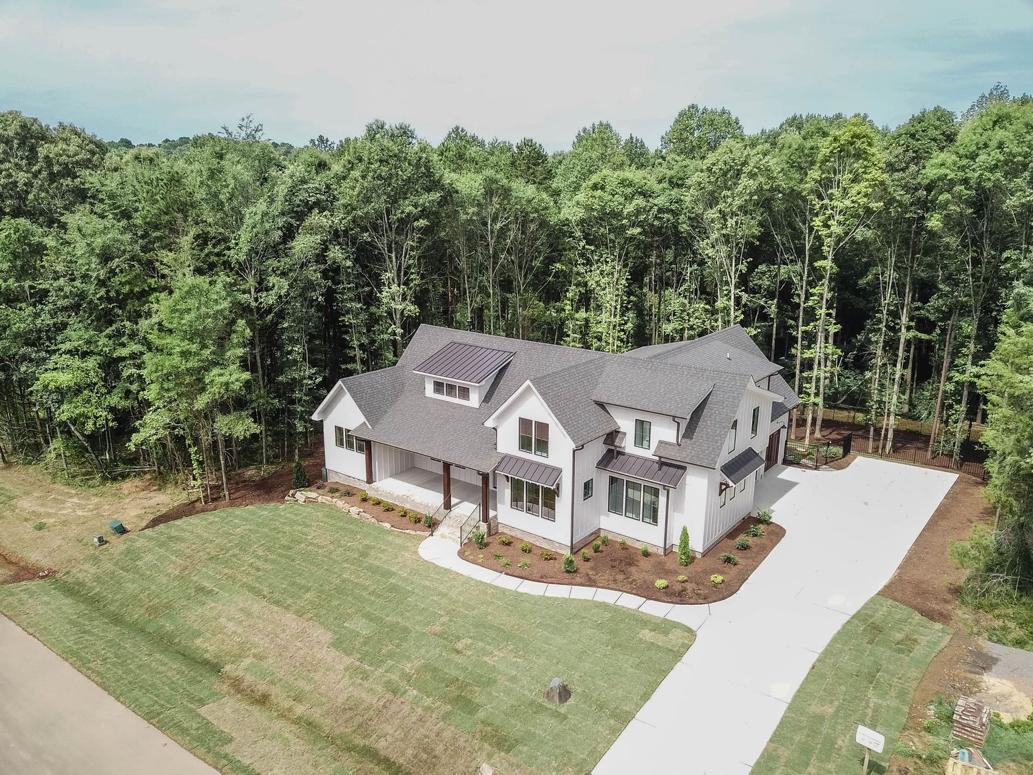Drone shot of elegant white farmhouse with attached garage nestled among trees.
