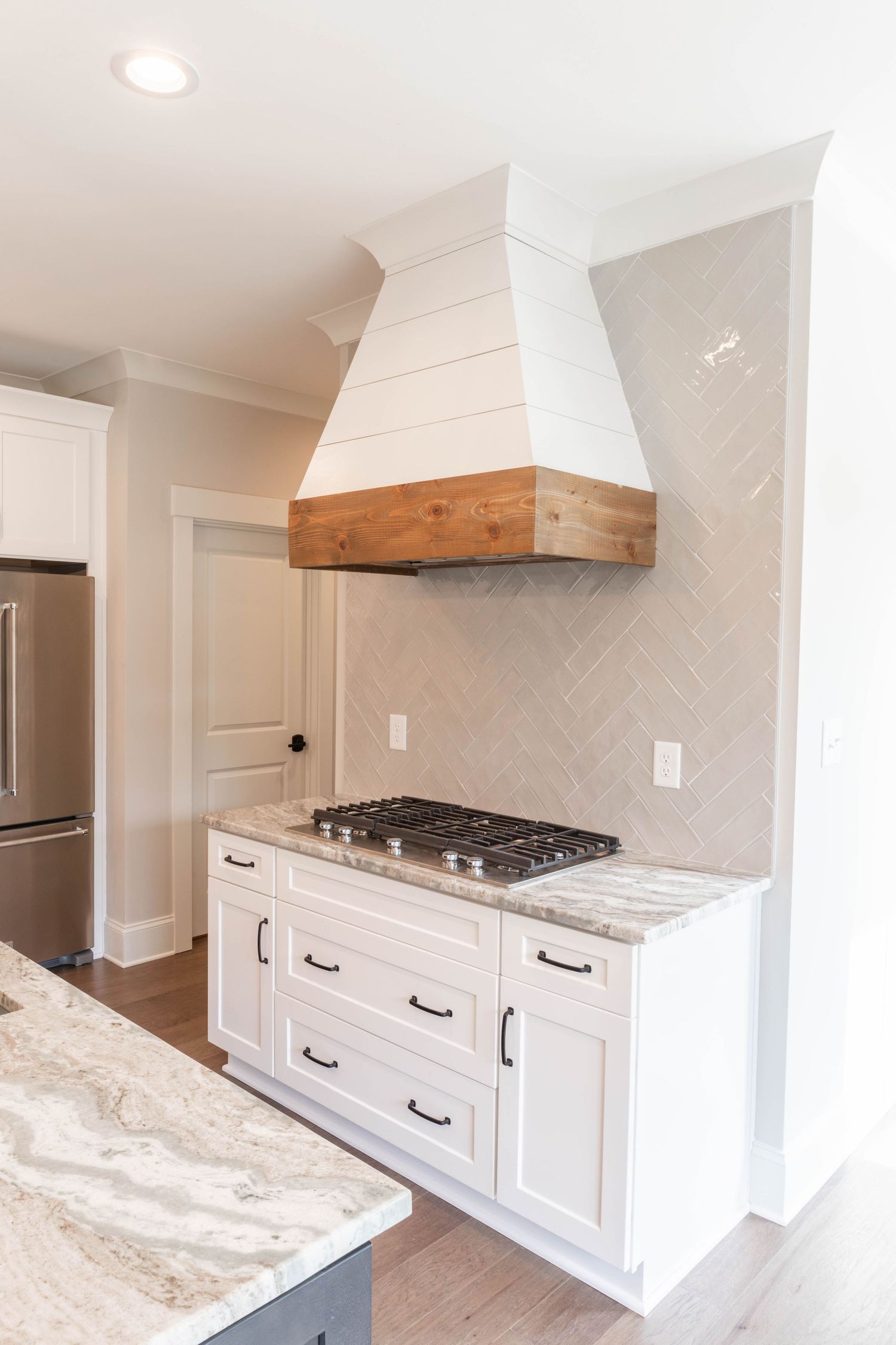 White kitchen cabinets with custom wood-trimmed range hood and herringbone tile backsplash.