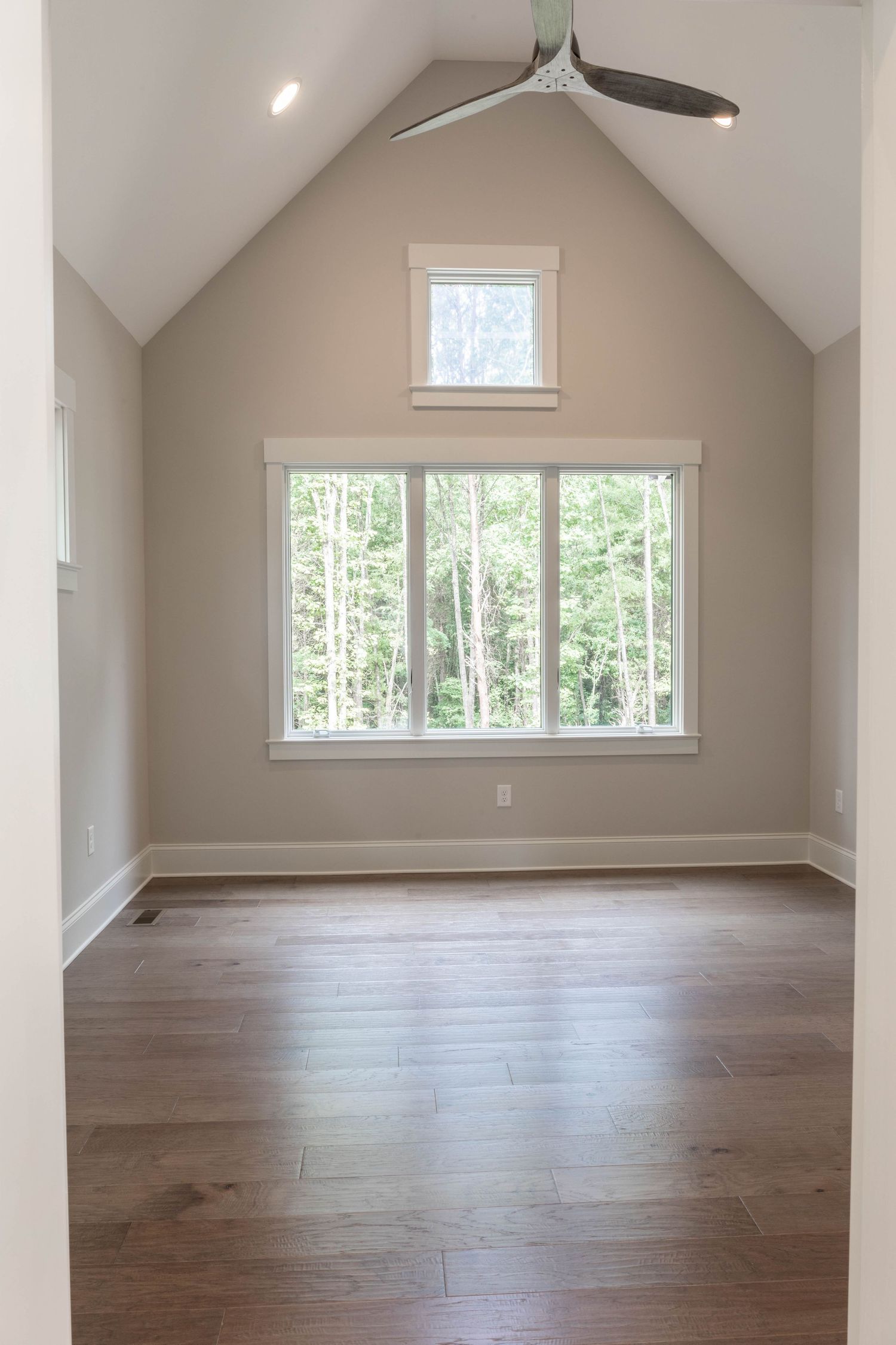 Vaulted ceiling room with modern ceiling fan and large windows overlooking forest scenery.