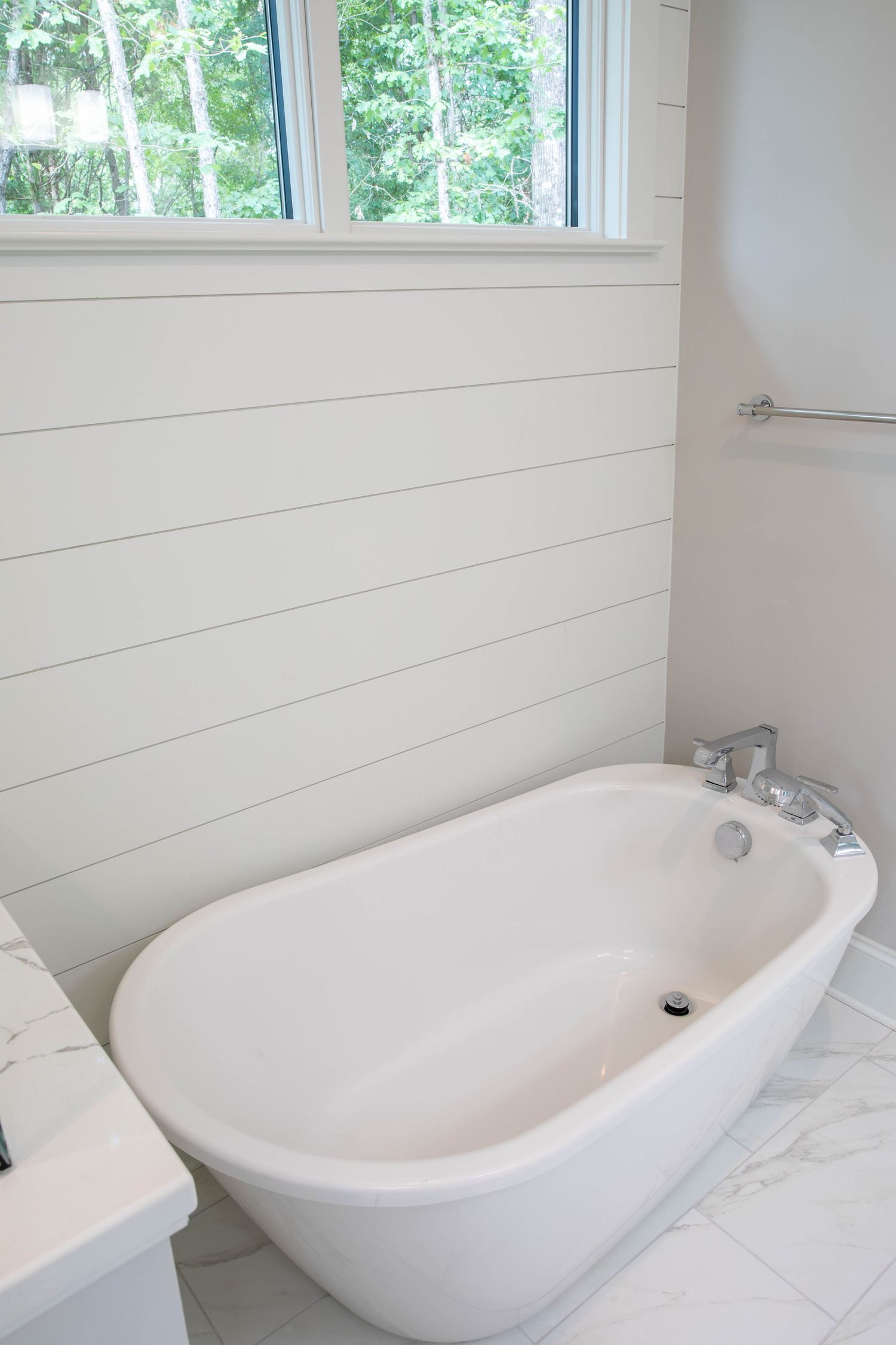 Modern freestanding white bathtub with shiplap walls and window overlooking trees.
