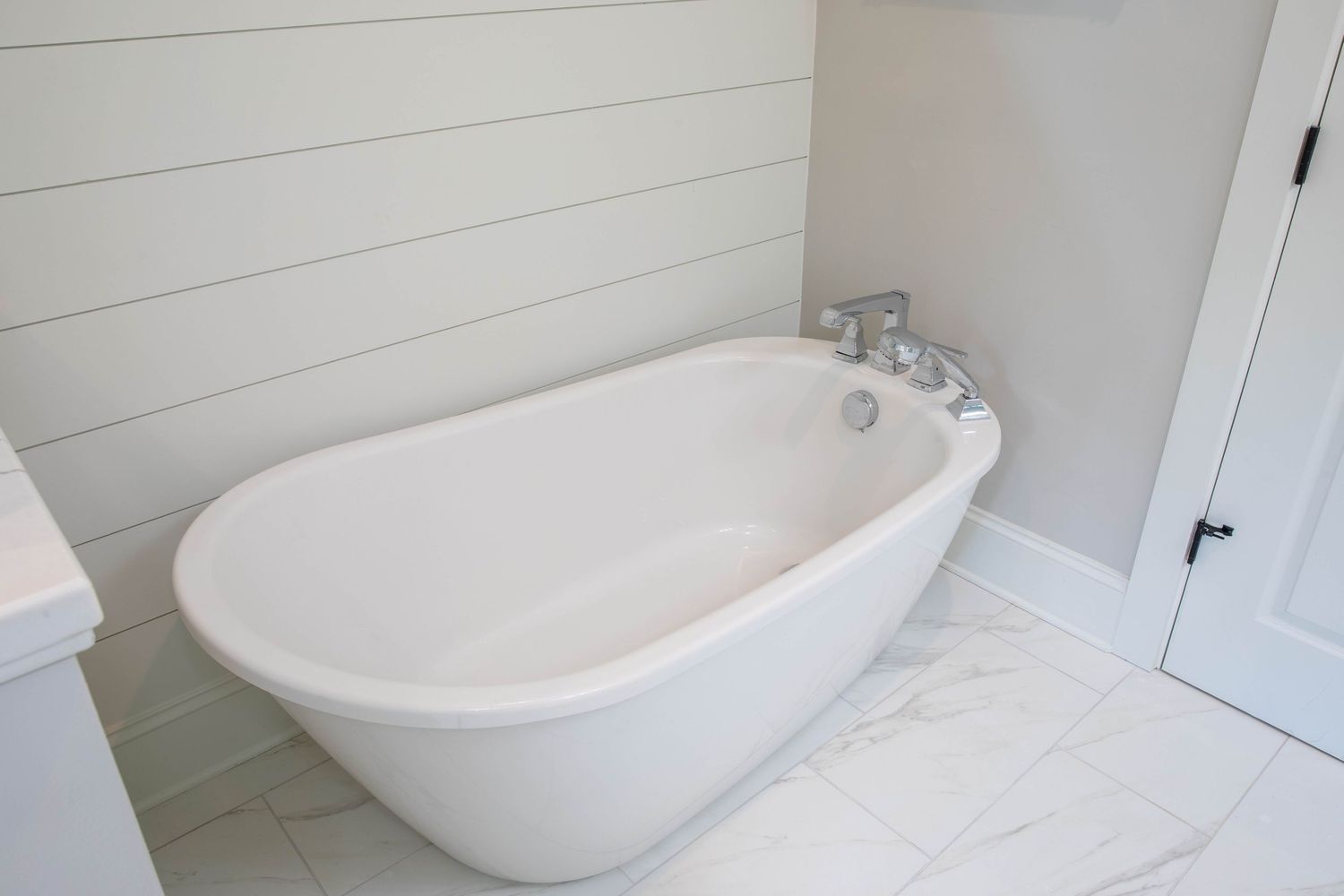 Elegant freestanding soaking tub in white bathroom with marble floors and shiplap accent wall.