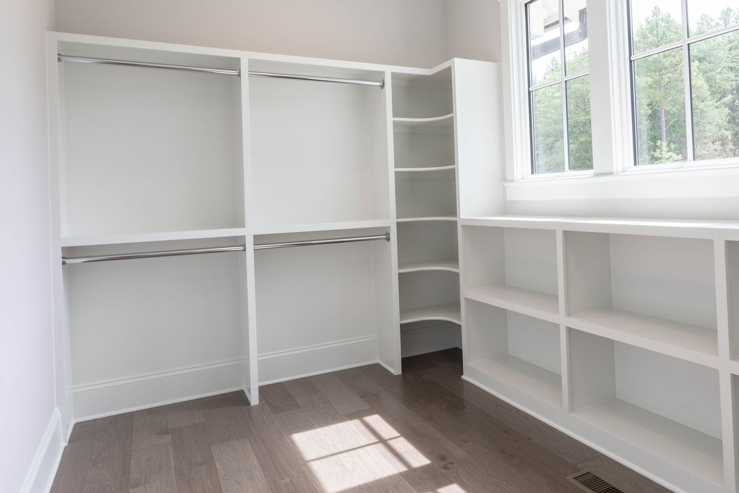 Walk-in closet with white shelving, hanging rods, and corner storage solution beneath windows.