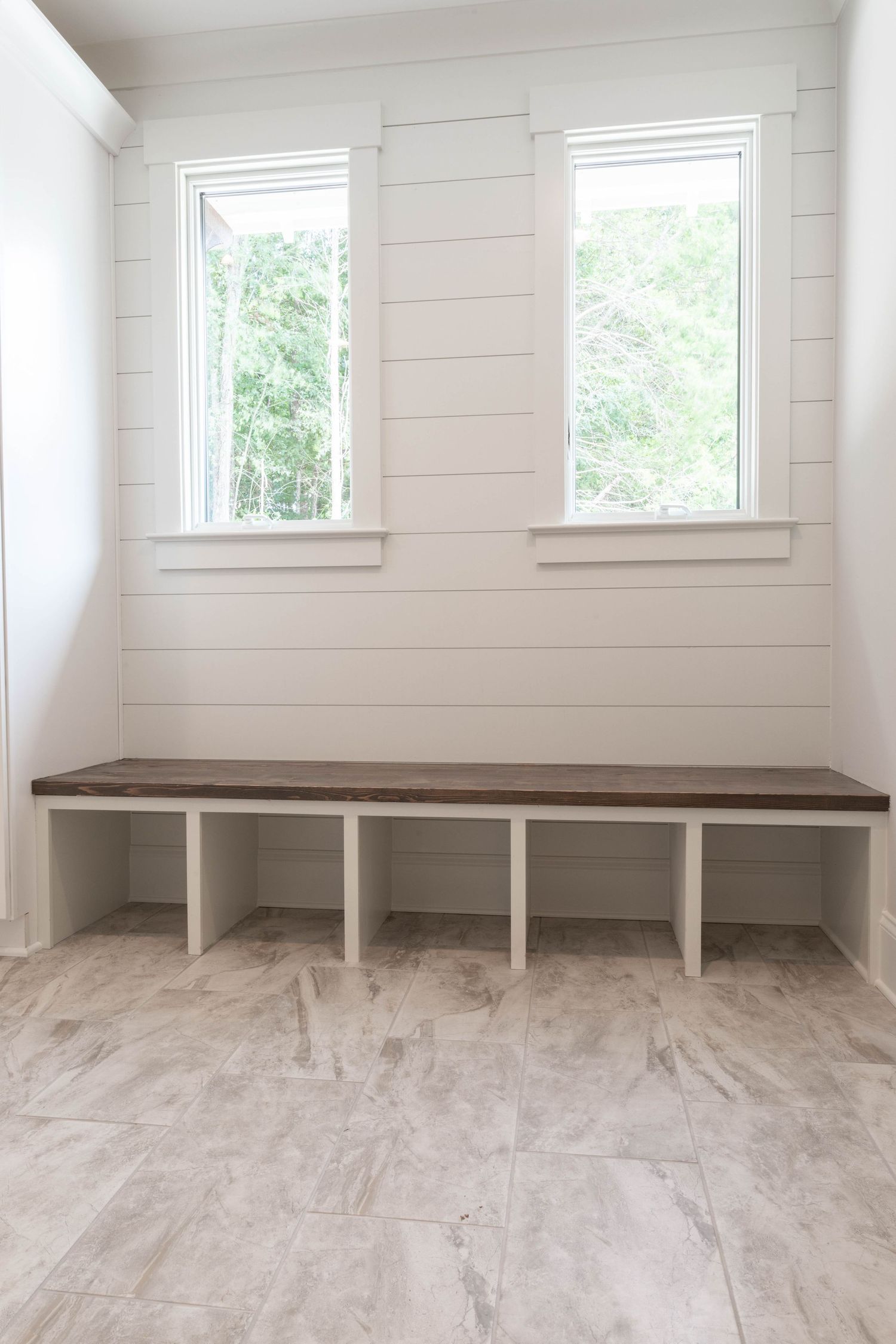 Modern mudroom with white shiplap walls, dual windows, and built-in wooden bench seating on grey marble-look flooring.