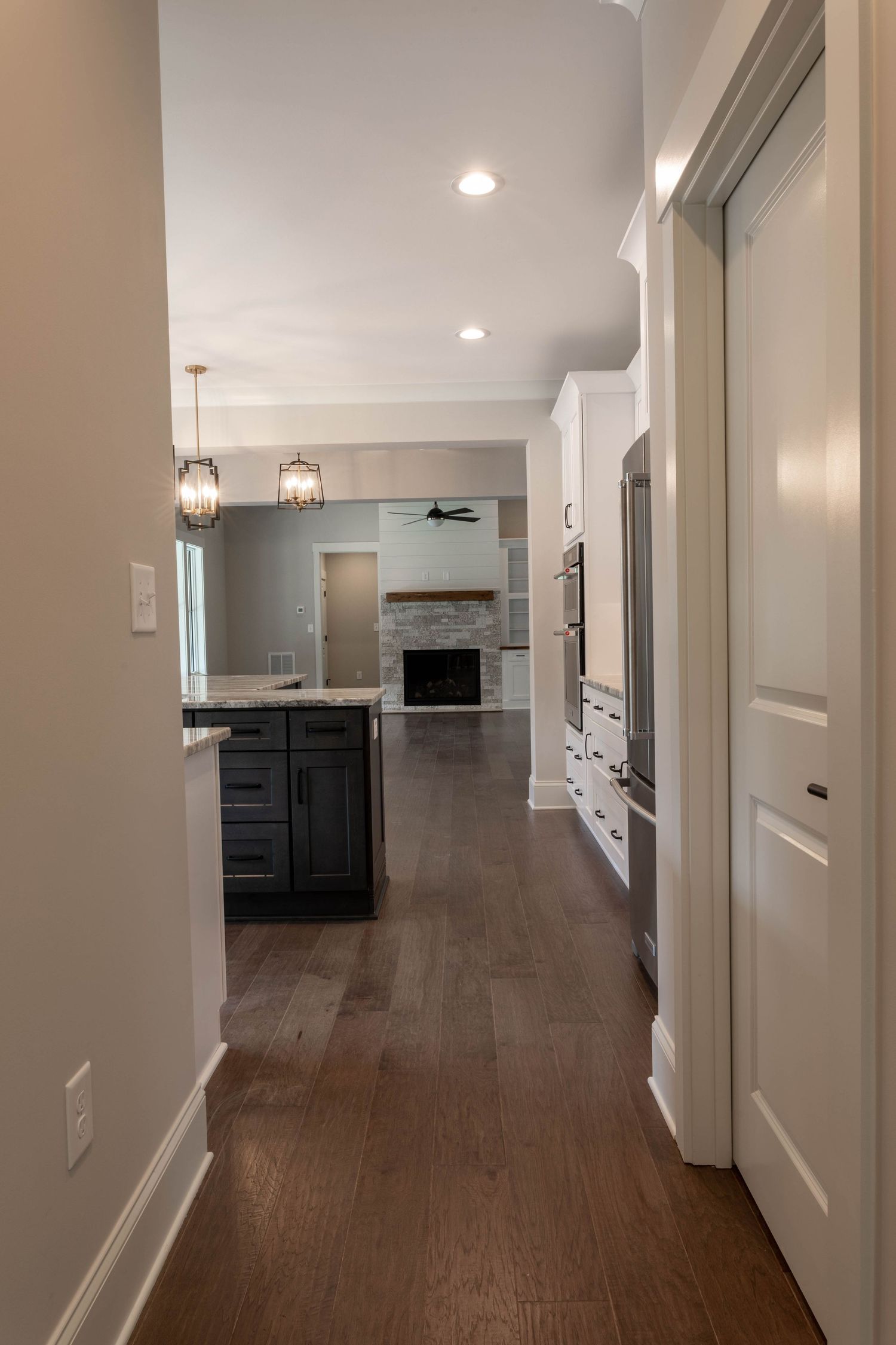 Modern hallway with hardwood floors leading to kitchen featuring pendant lights and dark cabinets.