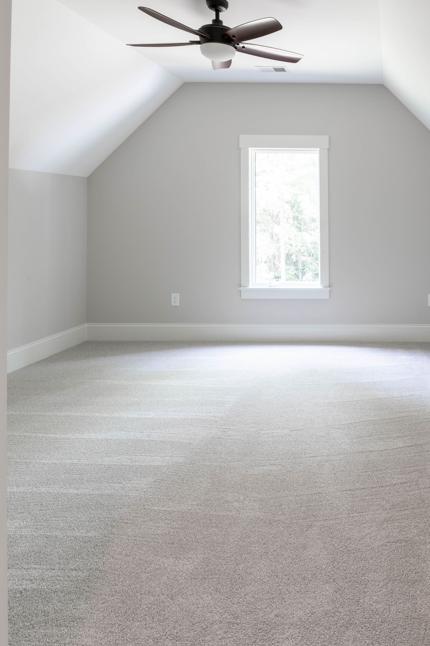 Spacious attic bedroom with wooden ceiling fan, window, and grey walls on light carpet.
