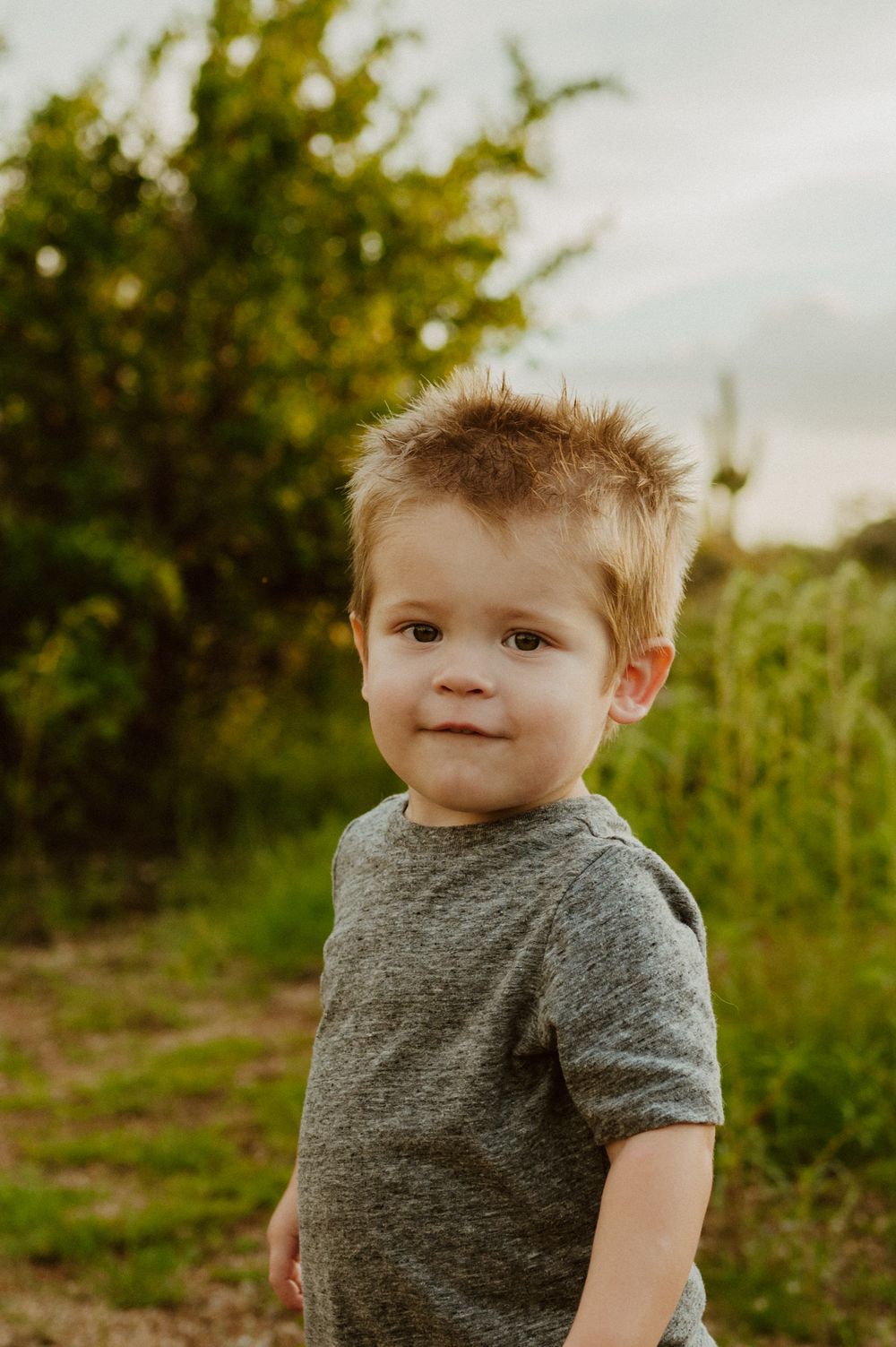 A toddler in a grey t-shirt smiles while outdoors during golden hour.
