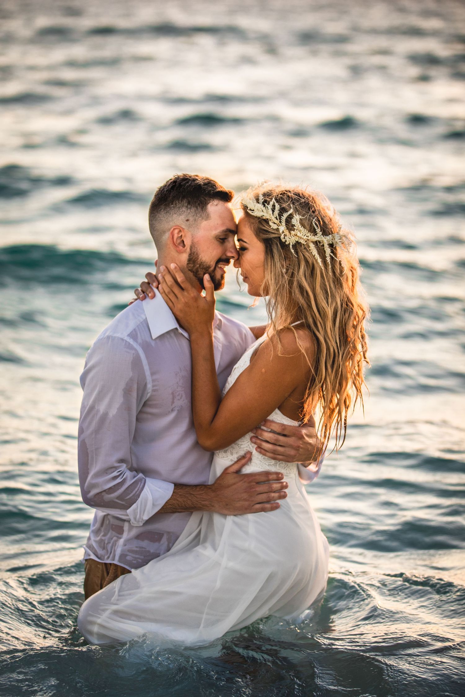 Romantic couple embracing in ocean waves during sunset photoshoot wearing white formal attire.