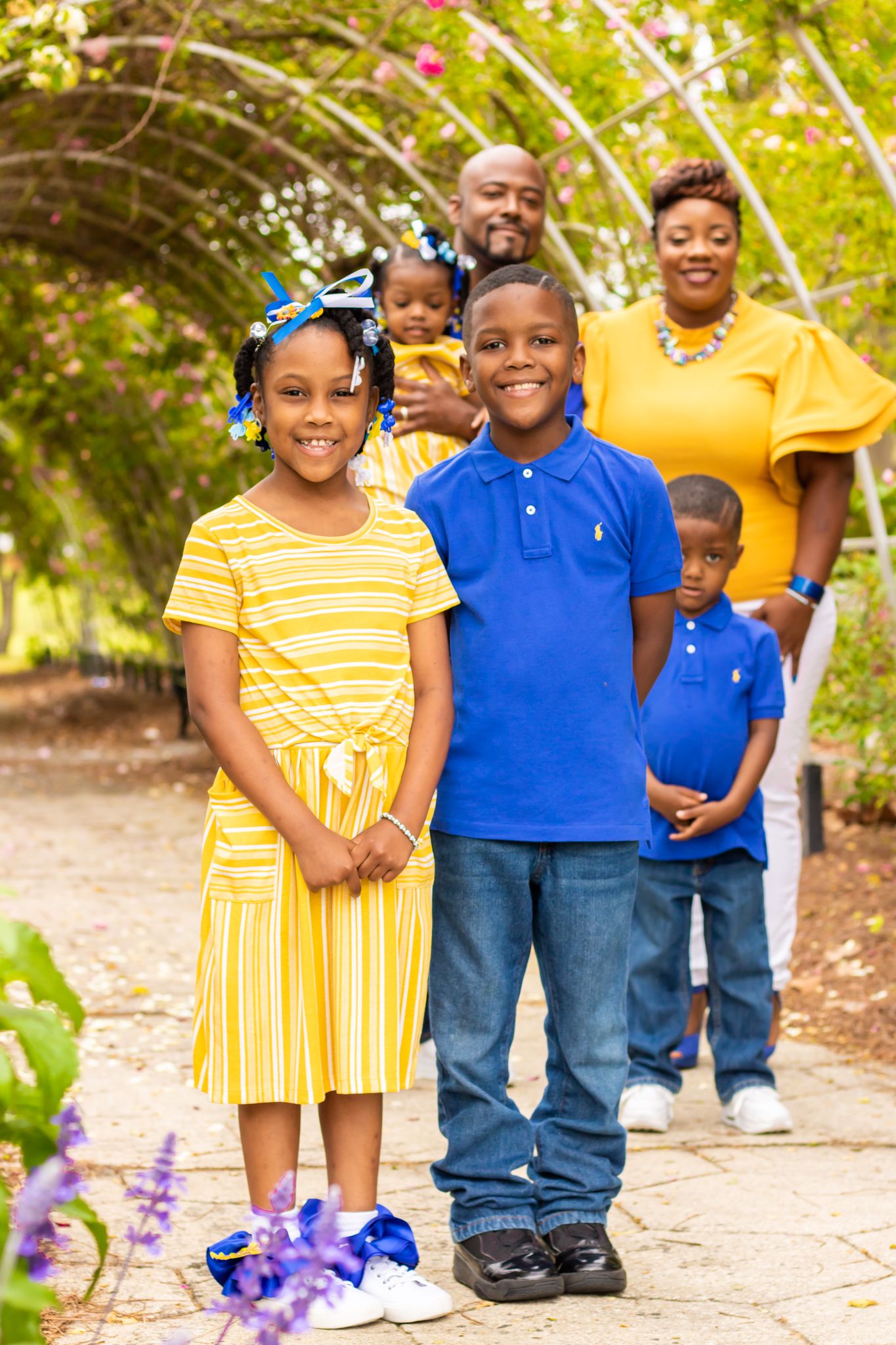 Family portrait under garden arch with children in coordinated yellow and blue outfits.