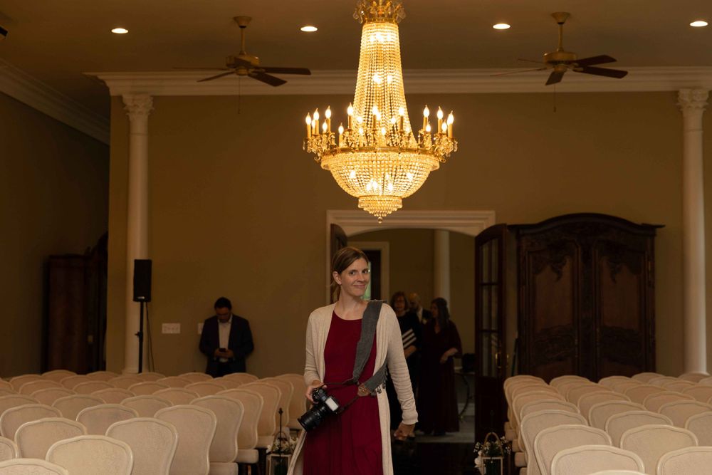 Photographer standing in elegant venue with crystal chandelier and rows of chairs.