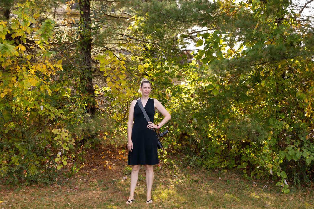 A silhouette poses outdoors against autumn foliage wearing a navy dress.