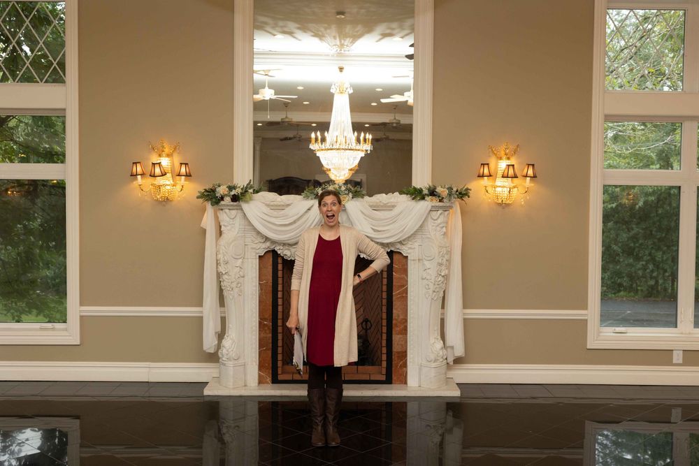 A figure stands in an elegant ballroom setting with crystal chandeliers and wall sconces beneath white fabric draping.