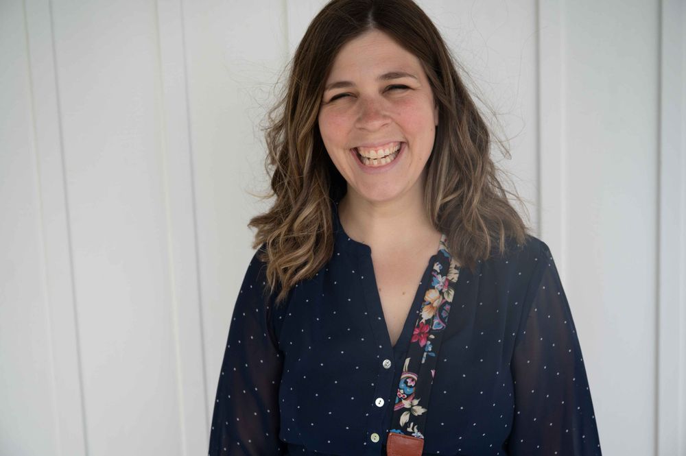 Someone wearing a navy polka dot blouse smiles warmly against a white paneled wall.