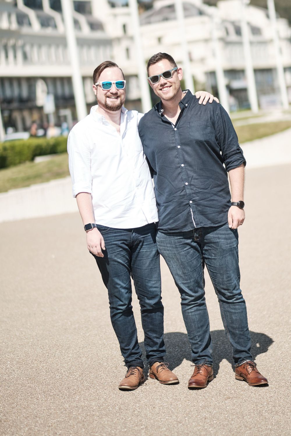 Two friends in casual attire stand together smiling on a sunny walkway.
