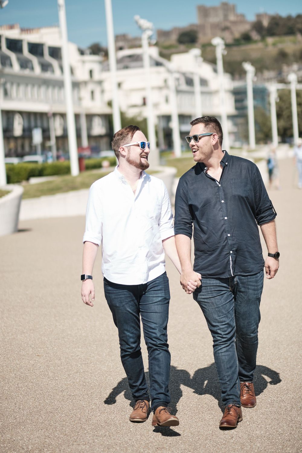 Couple walking hand in hand on a sunny promenade with white buildings in the background.