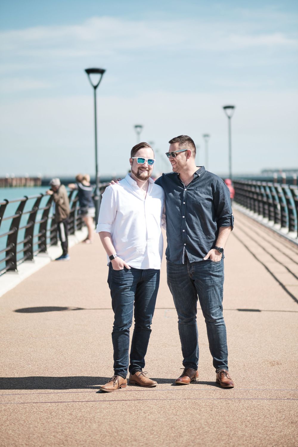 Two people wearing button-down shirts pose on a waterfront pier with lampposts in the background.