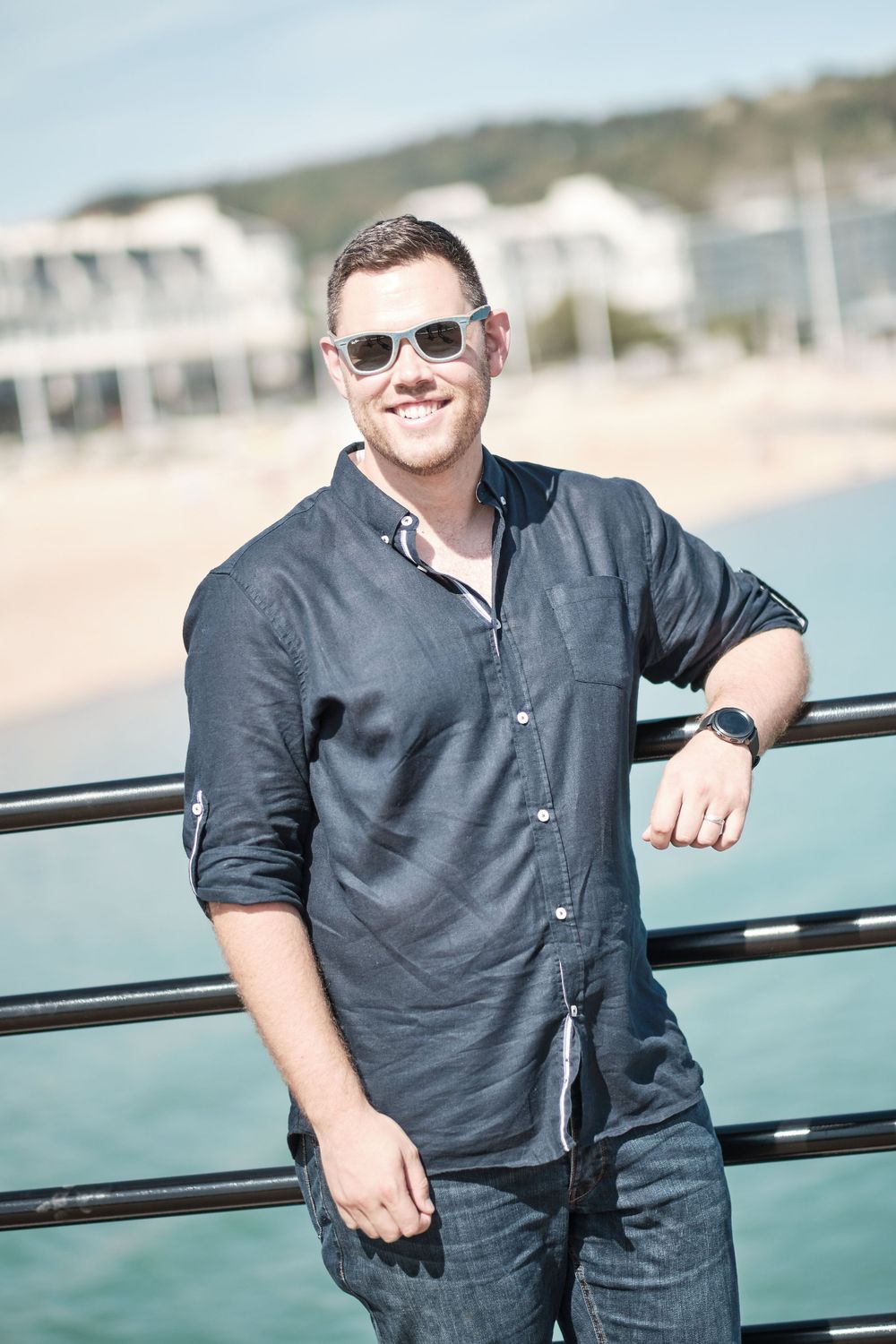 Person in dark shirt leaning against railing at waterfront location wearing sunglasses.