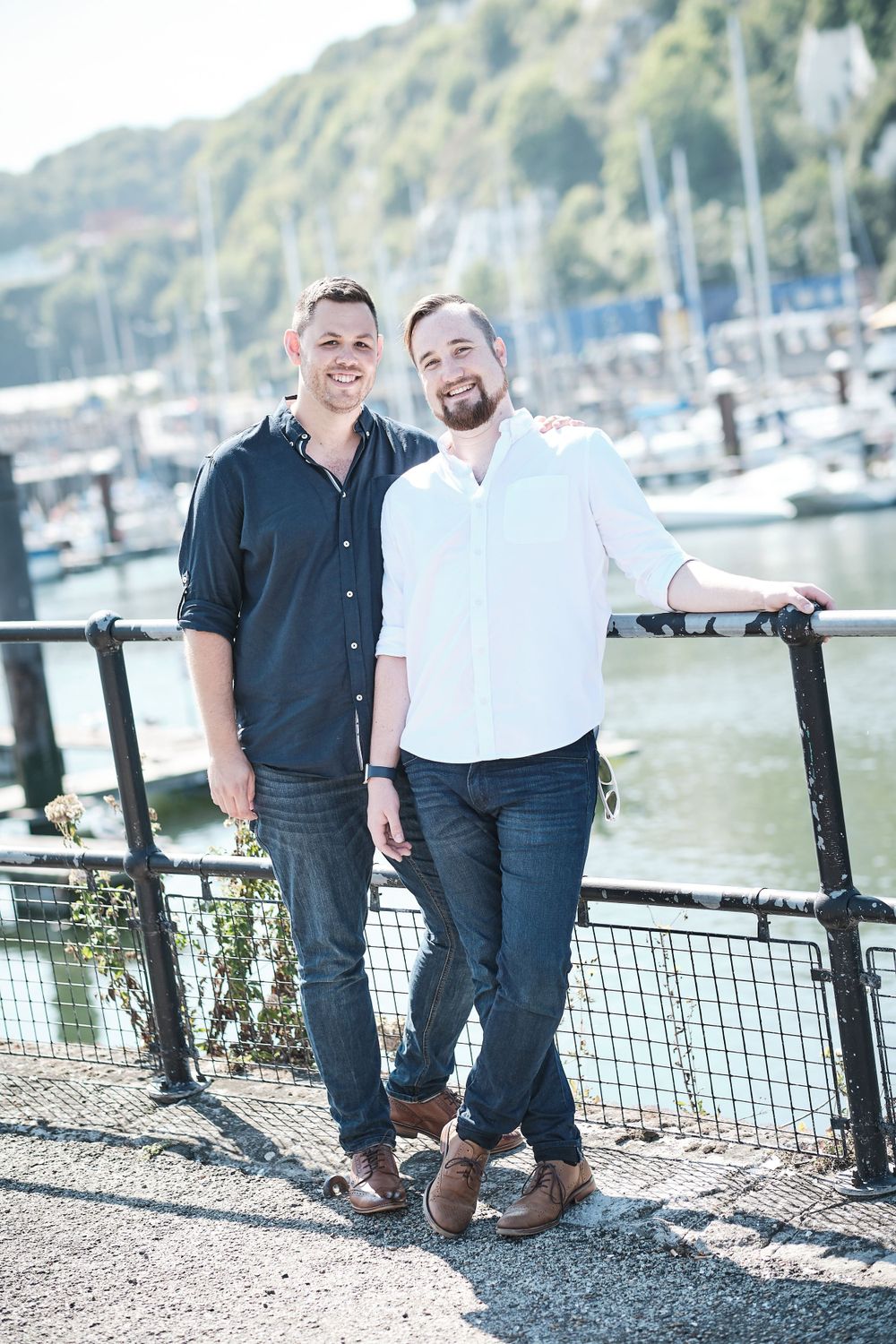 Two people in contrasting shirts pose together at marina with boats in background.