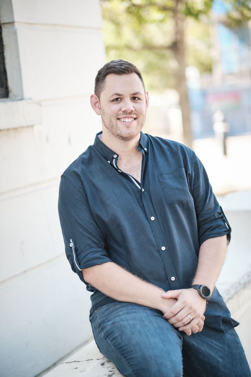 Person in navy button-down shirt sits casually against white wall outdoors.