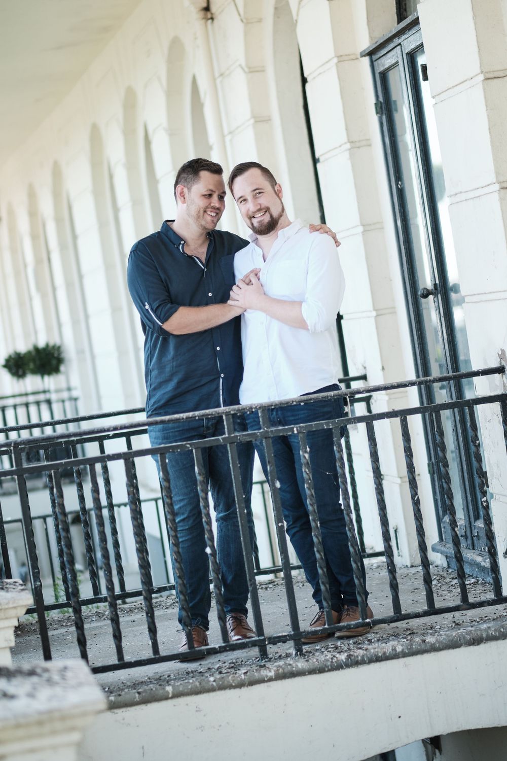 Two people in casual attire posing together on a balcony with white architectural columns.