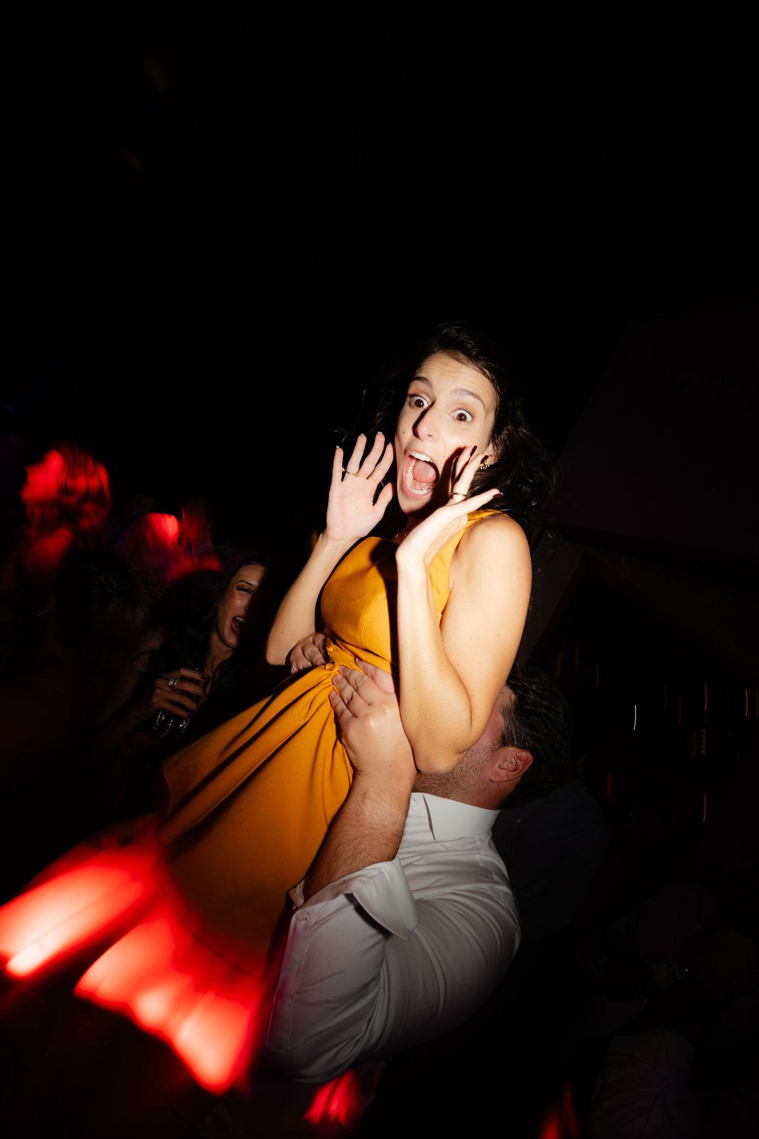 Person in yellow dress dancing enthusiastically in a dark room with red lighting.