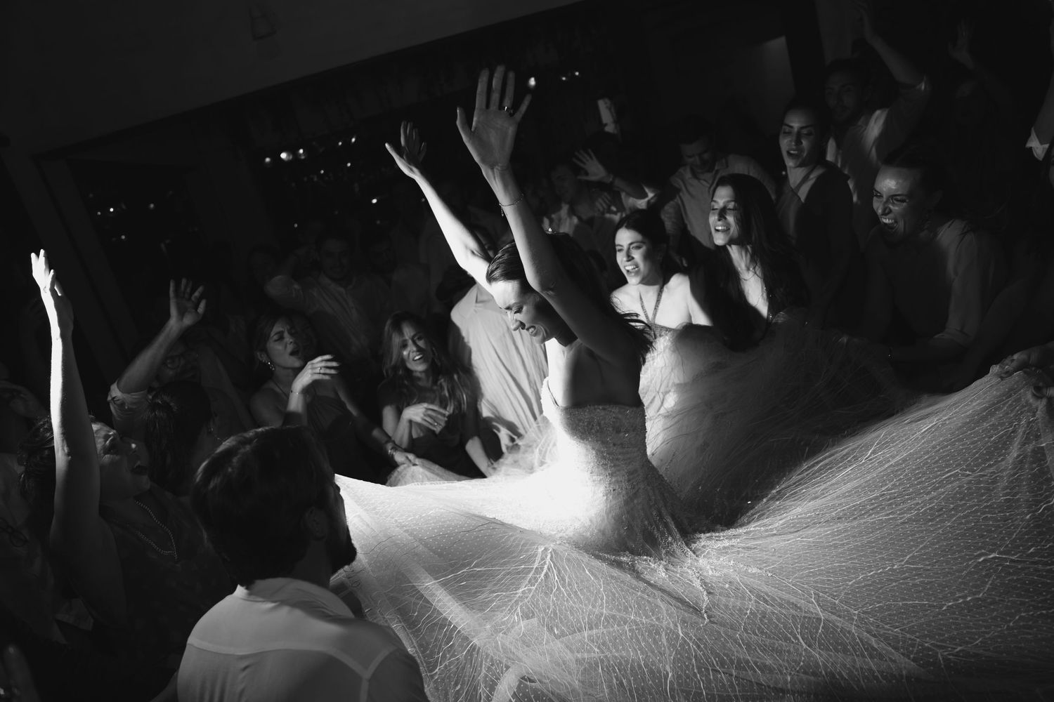 Black and white photo of wedding guests dancing around bride's flowing dress on dance floor.