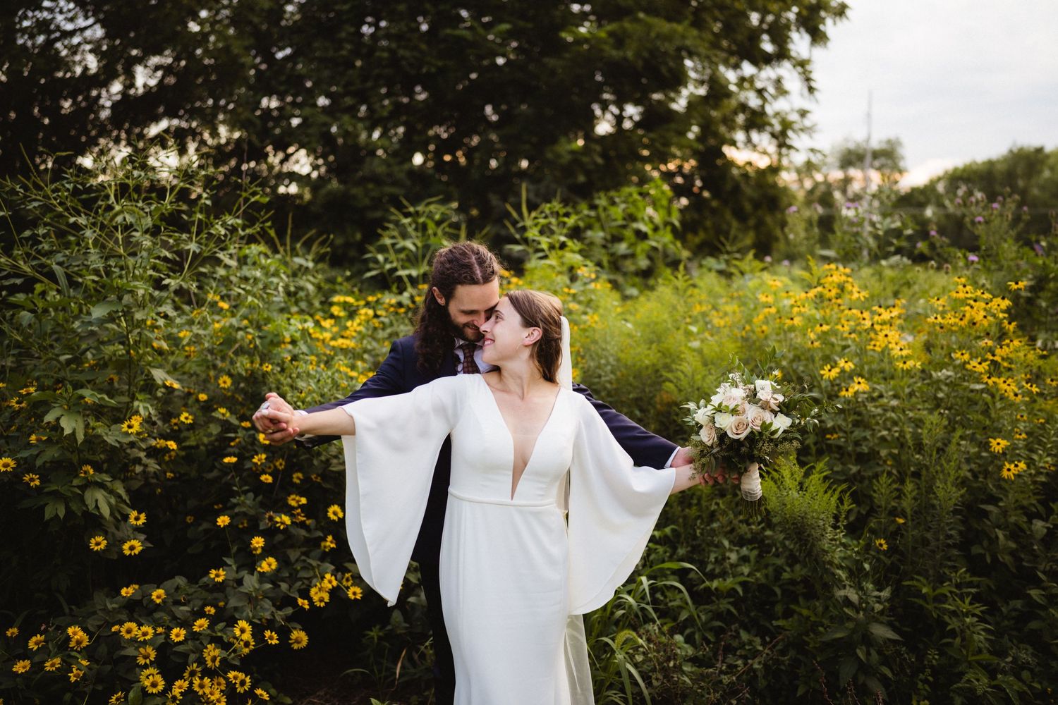 A bride in a flowing white dress with bell sleeves stands in a field of yellow wildflowers holding a bouquet.