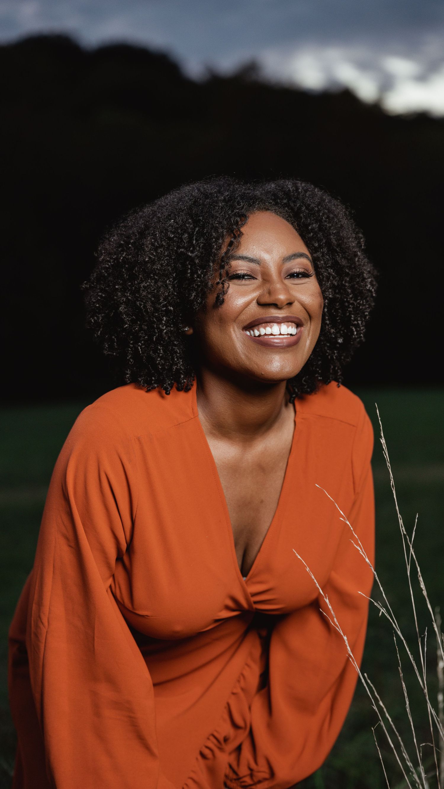 Smiling woman in orange dress laughing in field at dusk.