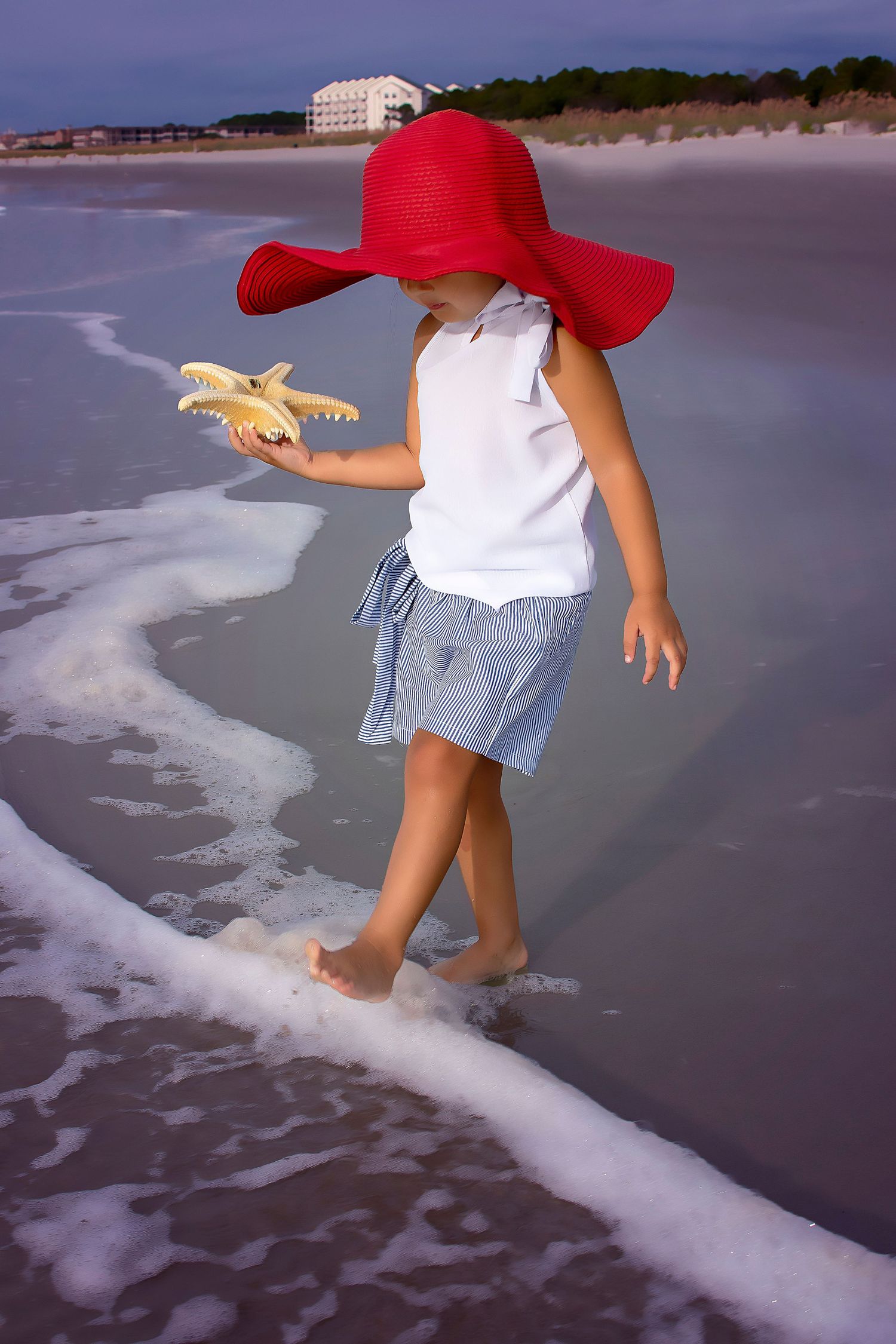 Child in red sun hat holds starfish while playing in ocean waves at the beach.