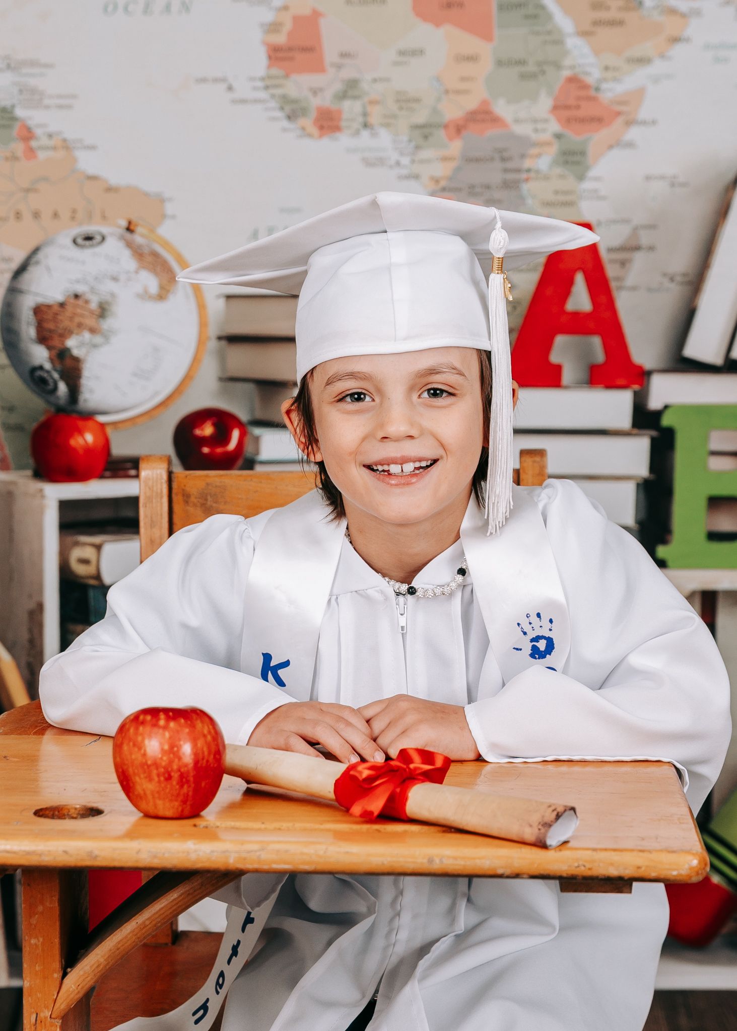 A smiling child in a white kindergarten graduation cap and gown sits at a desk with an apple and diploma.