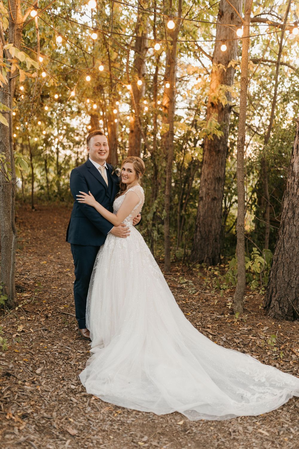 Bride and groom posing together under string lights among tall trees at golden hour.