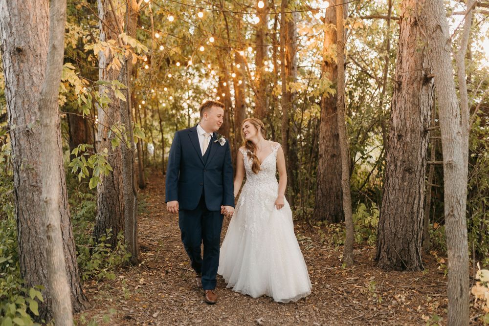 Bride and groom walking hand in hand through sunlit forest path in autumn.