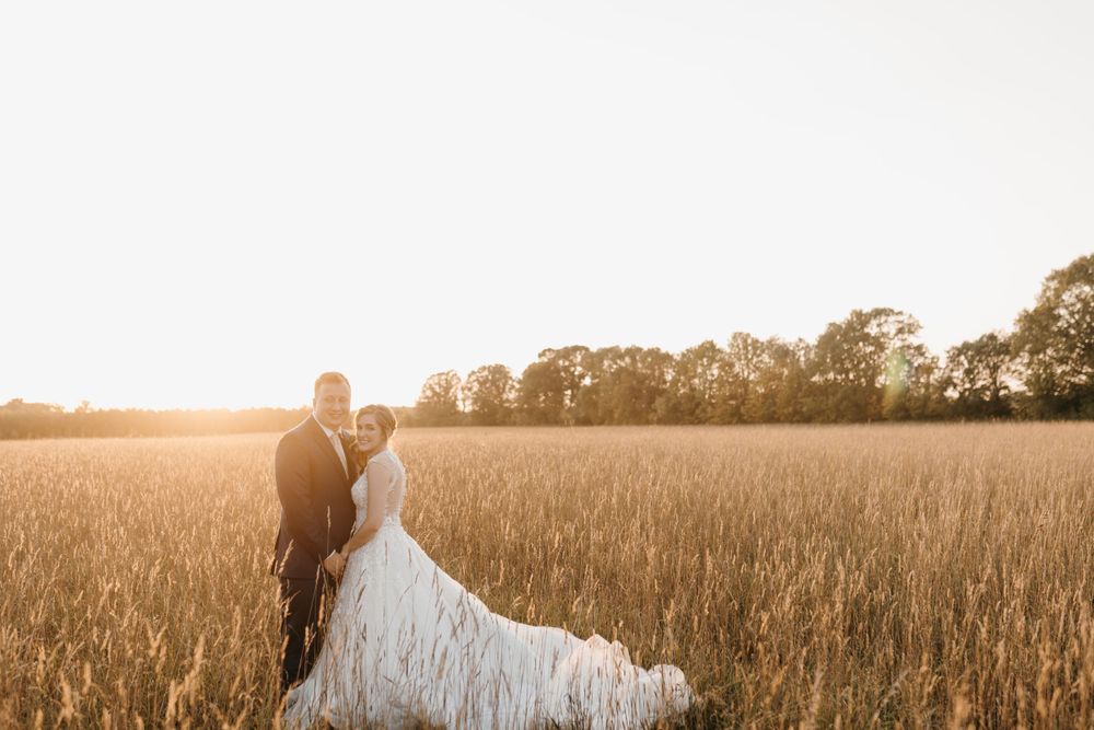 Bride and groom stand in golden wheat field at sunset, brides long train flowing gracefully behind her.