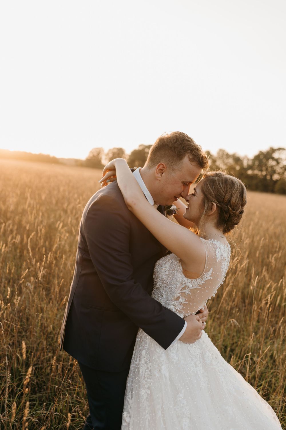 Groom tenderly holds bride in lace gown as they share an intimate moment in a golden sunset field.